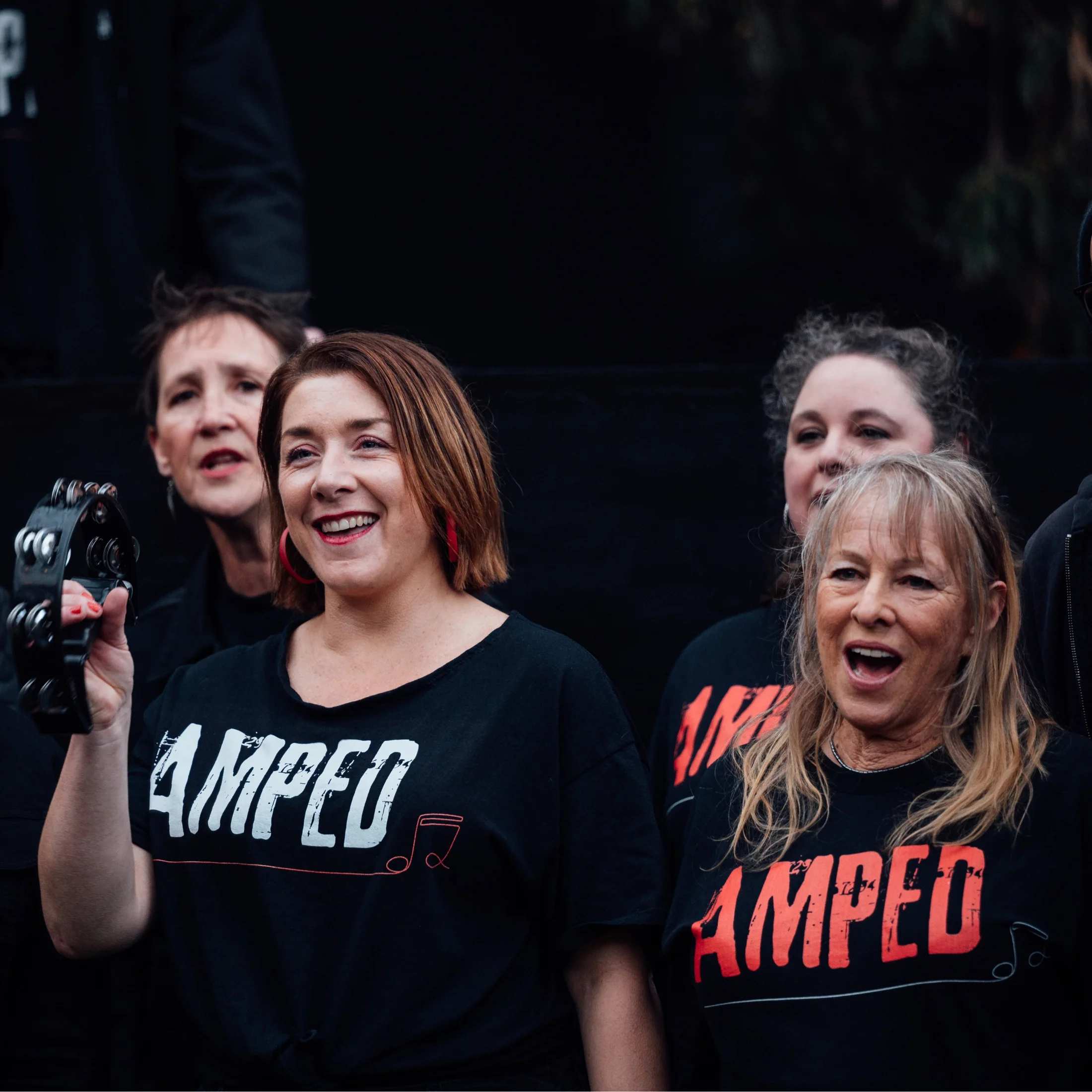 A colour photograph of people singing in an outdoor setting. They're wearing black t-shirts with the text 'amped' written across the front. The event is a part of the Festival of Voices program