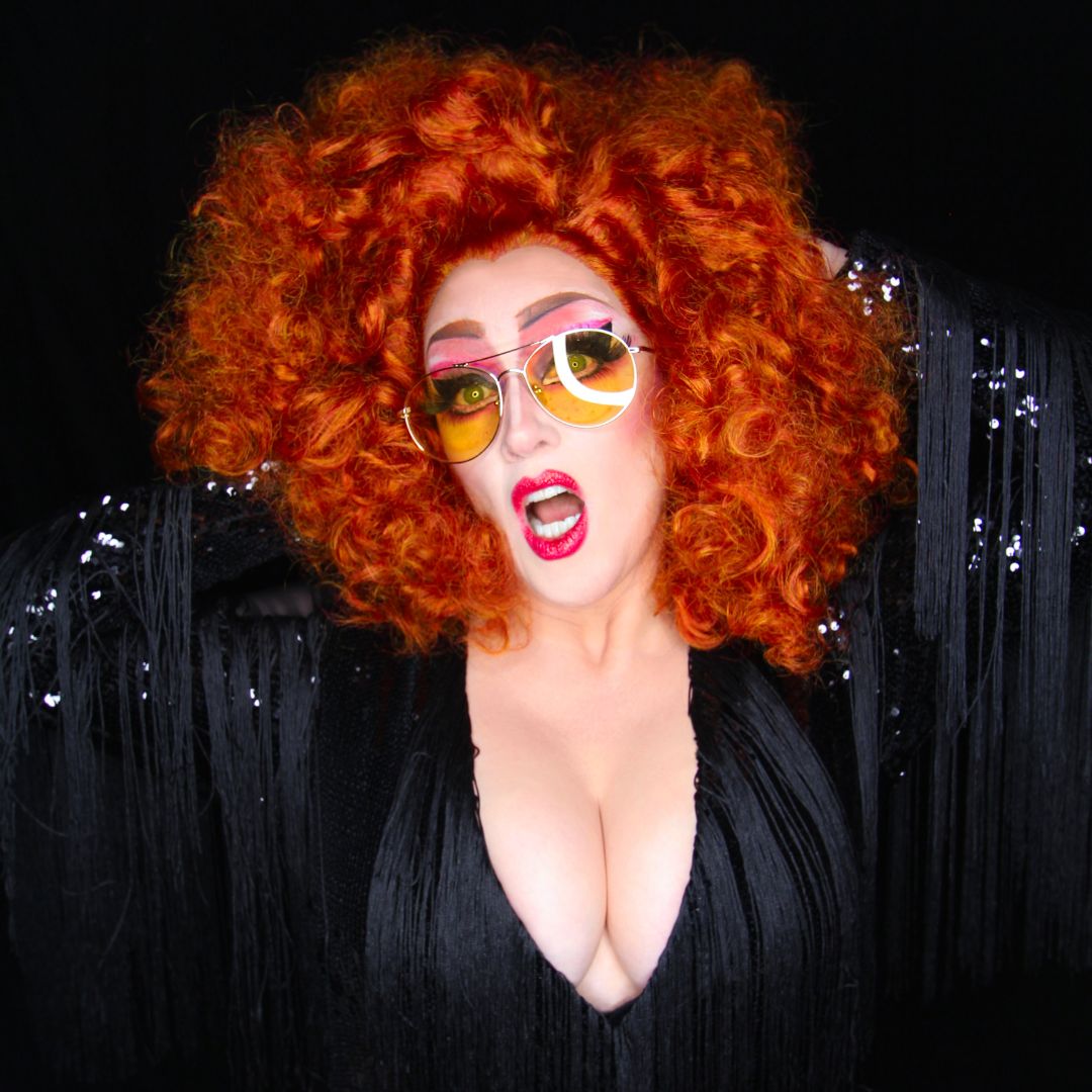 A photograph of a woman with orange hair and bright makeup wearing yellow sunglasses, dressed in black and in front of a black sparkly backdrop.