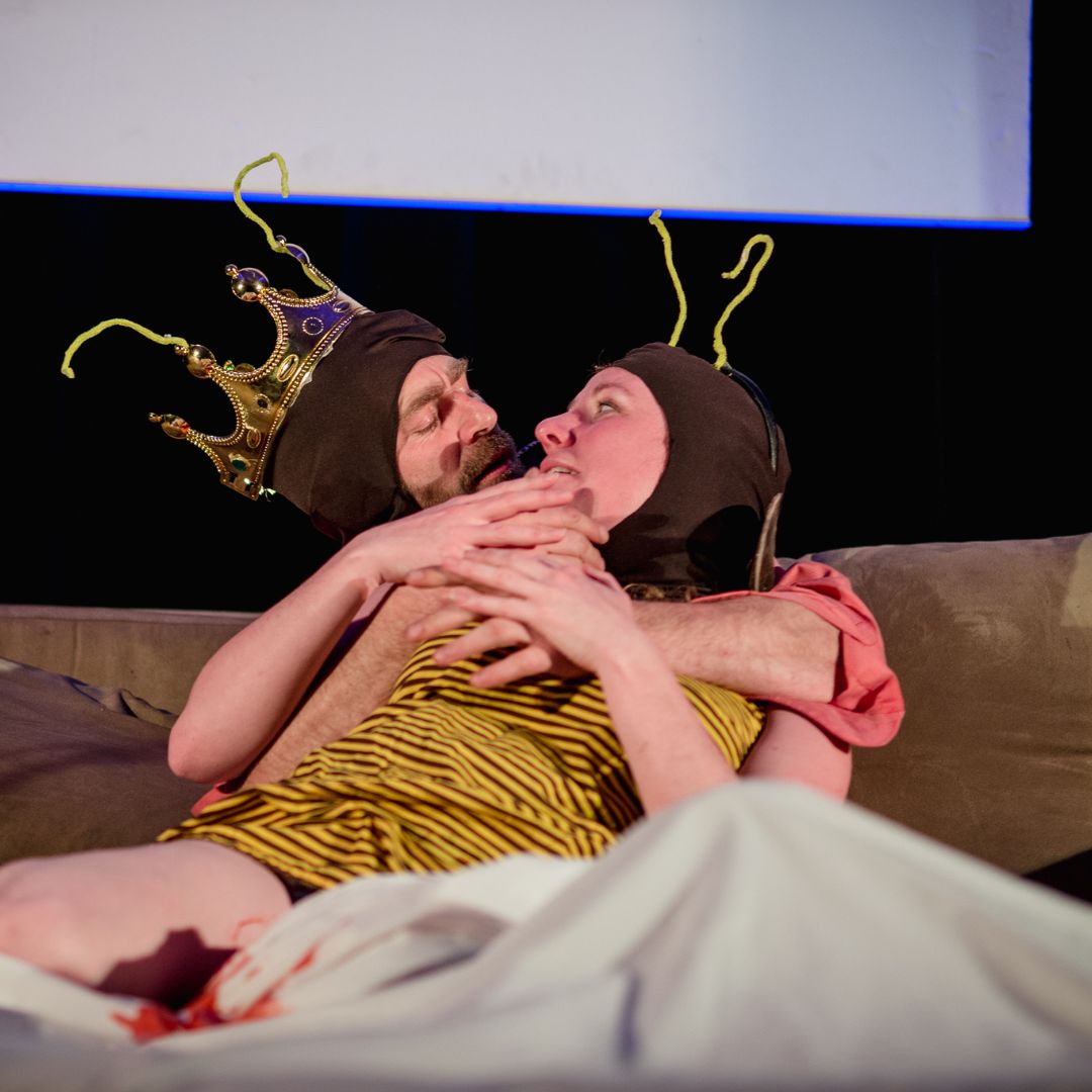 Two people are embracing, in costume, seated on a couch on stage