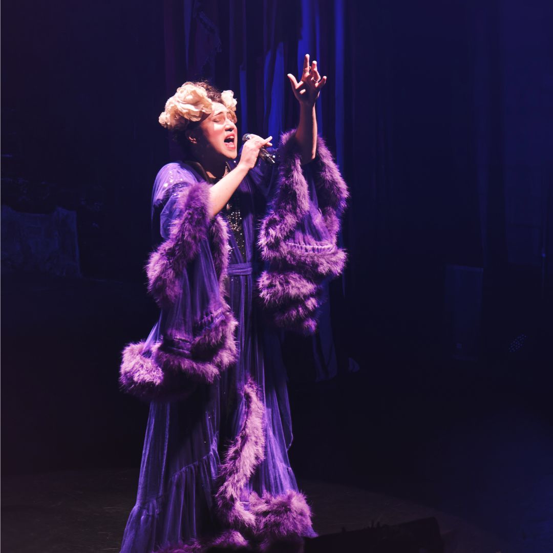 A photograph of a woman dressed in purple stage costume singing on stage in front of a dark backdrop