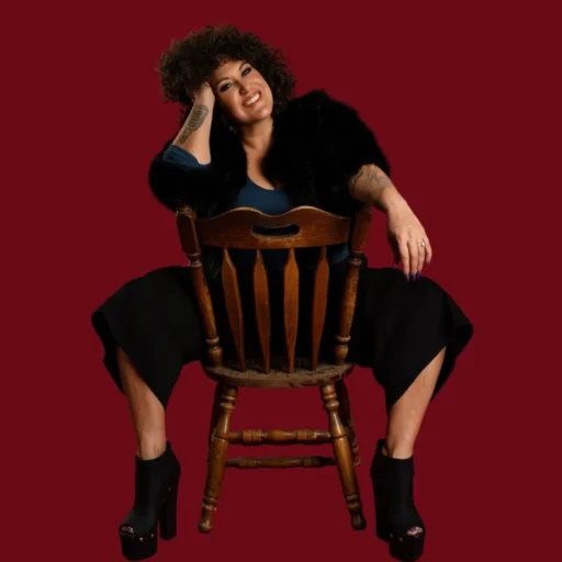 Casey Donovan; a woman with curly hair sits confidently on a wooden chair turned backward, smiling warmly at the camera. She wears a dark top, black fur-style jacket, wide-leg pants, and high platform heels against a deep red background.