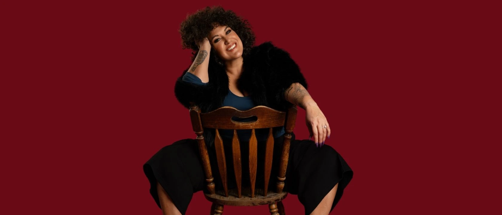 Casey Donovan; a woman with curly hair sits confidently on a wooden chair turned backward, smiling warmly at the camera. She wears a dark top, black fur-style jacket, wide-leg pants, and high platform heels against a deep red background.
