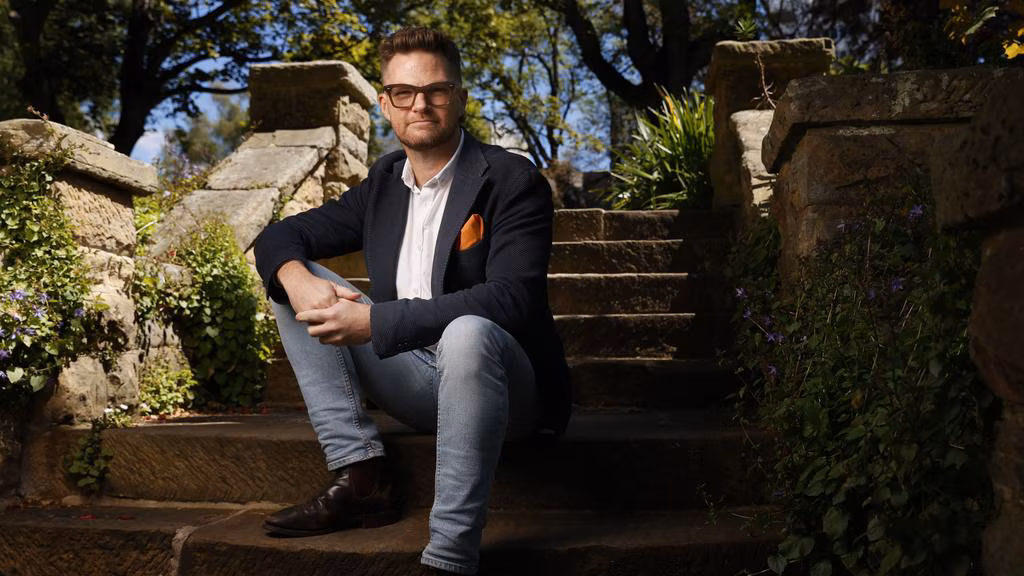 A man identified as Shaun Rennie, wearing a dark blazer, light blue jeans, and an orange pocket square, sits on an outdoor stone staircase surrounded by green foliage, looking directly at the camera.