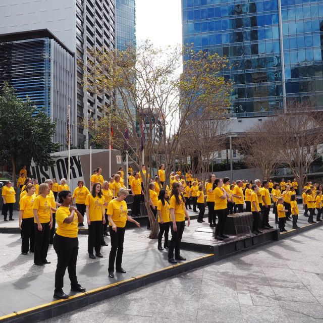 a large group of people singing together as a choir wearing yellow t-shirts outdoors in a city background